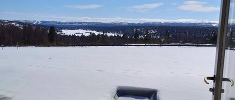 Vista desde una habitación con terraza de madera que da a un paisaje nevado, que incluye un pequeño jardín y una estructura metálica.
