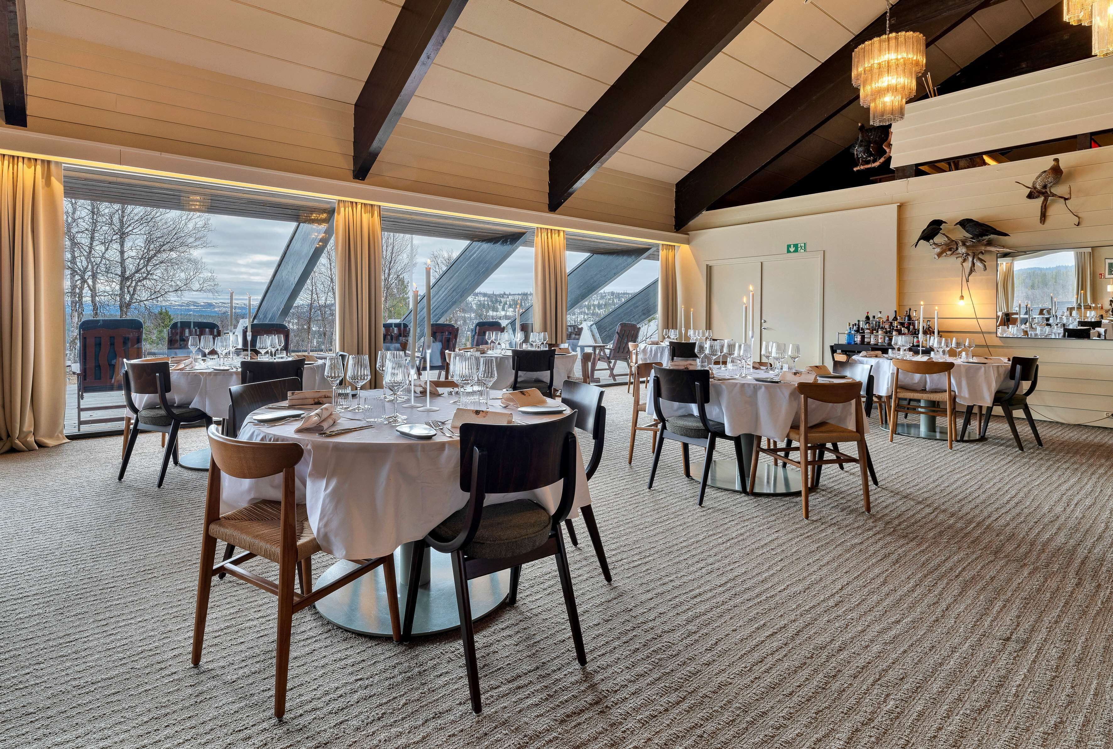 A restaurant with multiple round tables set for dining, large windows with curtains, and a chandelier.