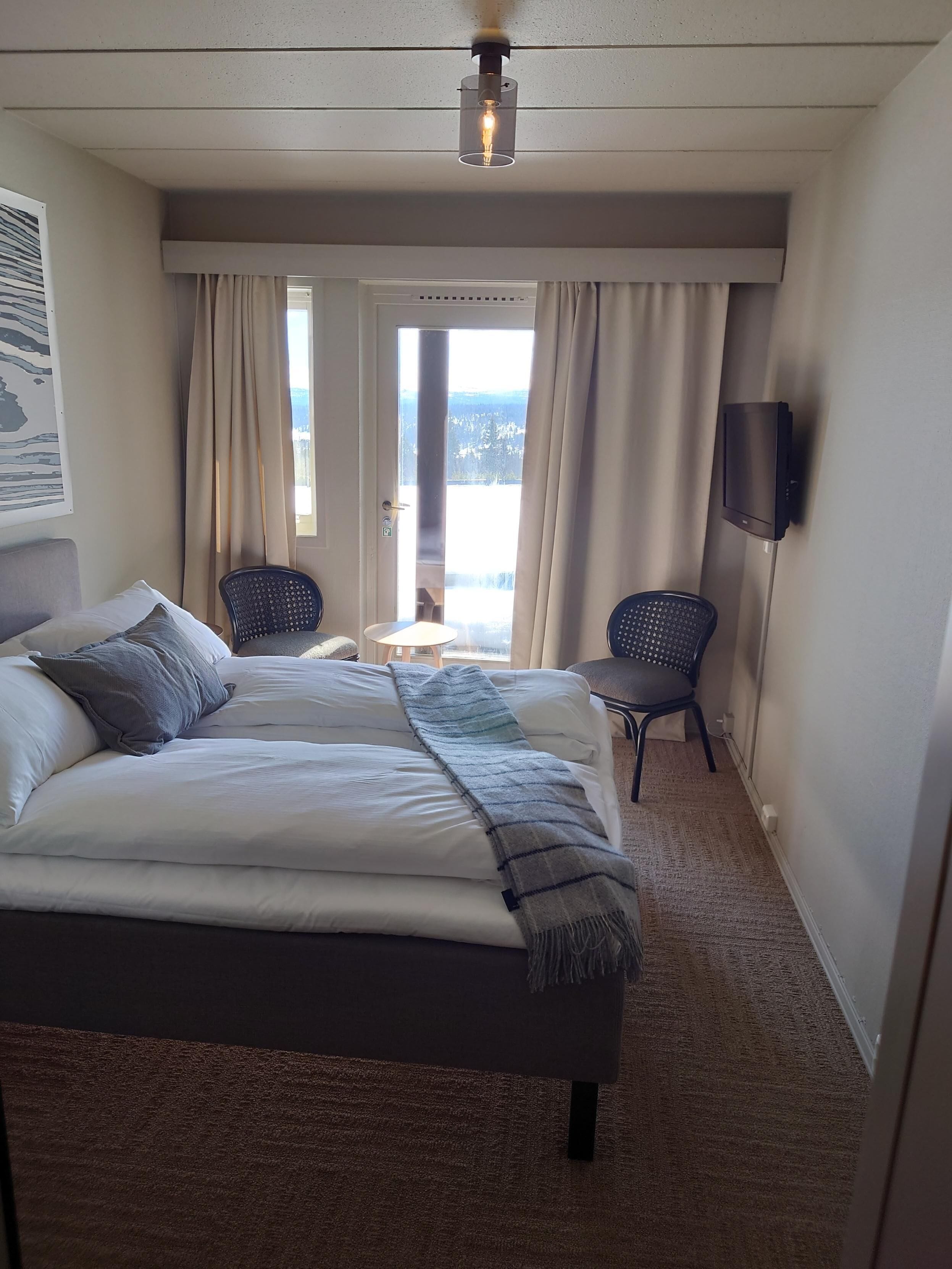A hotel room with a large bed, two chairs, a TV, and a window with curtains.