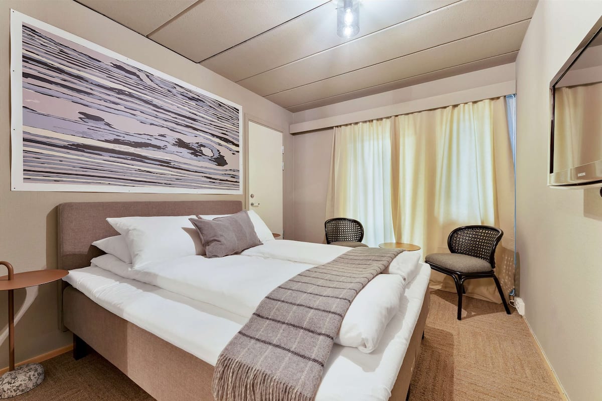 A hotel room with a bed, a chair, a small table, a painting on the wall, and a window with curtains.