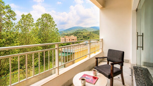 A balcony with a view of green hills and a building in the distance. There is a chair and a small table with a cup and a book. - Mewar Palace Luxury Resort & Private Pool Villas - Udaipur (Udaipur)