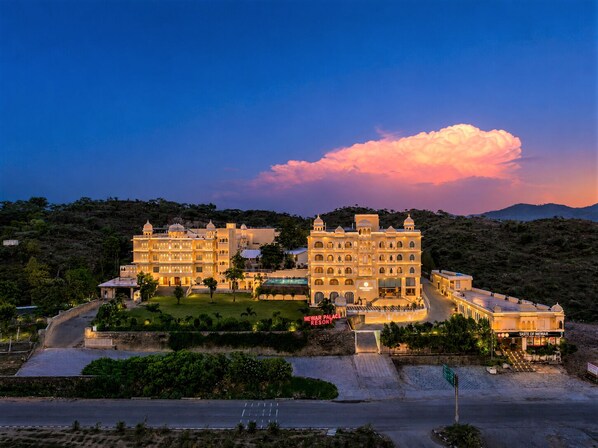 A large, multi-story building with a central courtyard, surrounded by lush greenery and hills in the background.