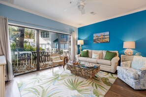 A living room with a large sliding glass door leading to a balcony, a ceiling fan, a painting on the wall, and a patterned rug. - 47 Ocean Breeze: Pet Friendly 2 BR Villa Next to Pool, Walk to Beach (Hilton Head Island)