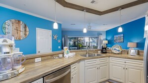 Une cuisine avec un plan de travail en bois, des armoires blanches, un lave-vaisselle encastré et un mur bleu orné d’une horloge et d’objets décoratifs.