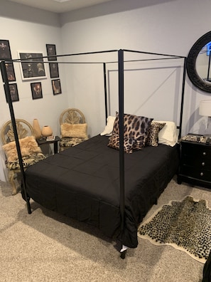 Una cama con dosel con un edredón negro, almohadas estampadas de leopardo y una alfombra a juego. Hay cuadros en la pared, un espejo redondo y una mesita con una lámpara.