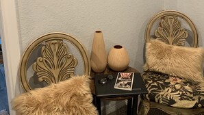 Ein Zimmer mit einer weißen, strukturierten Wand, einem kleinen Tisch mit einer Vase, zwei Stühlen mit Fellkissen und mehreren gerahmten Kunstwerken.