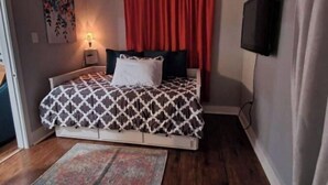 A bedroom with a bed, a nightstand, a wall-mounted TV, a red curtain, and a floral painting. - Location! Location! Location!  (Houston)