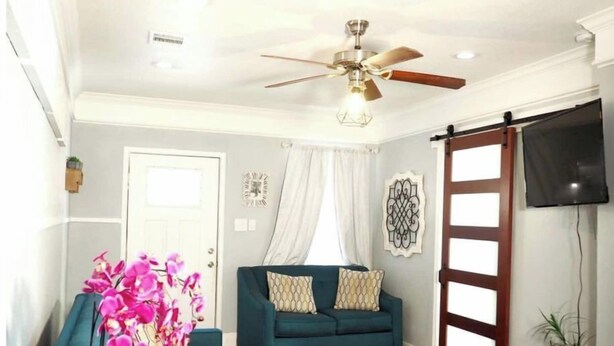 A living room with a teal sofa, a ceiling fan, a door with a curtain, and a wall-mounted TV. - Location! Location! Location!  (Houston)