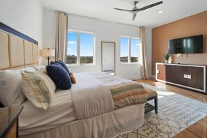 A bedroom with a large bed, a ceiling fan, a TV, and a view of the outdoors.