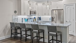A modern kitchen with white cabinets, a stainless steel refrigerator, a built-in microwave, and a marble island with bar stools. - Villa #706: 4  BR, 3  BA Condominium in Garden City, Sleeps 8 (Garden City)