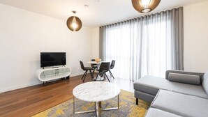 Una sala de estar moderna con un sofá, un televisor en un mueble de vidrio, una mesa de comedor con sillas y un ventanal con cortinas.