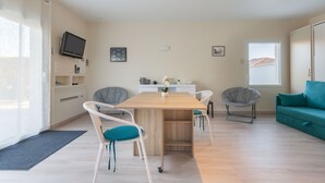 A modern living room with a wooden dining table, white and grey chairs, a teal sofa, and a television mounted on the wall.