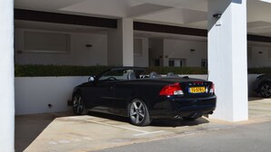 Um carro conversível preto estacionado numa área de estacionamento coberta, com paredes brancas e um hedge verde.