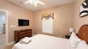 Une chambre à coucher comprenant un lit, une commode, un téléviseur, une fenêtre avec des stores et un ventilateur de plafond.