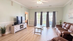A living room with a TV on a wooden stand, a brown leather couch, a coffee table, and a view through large windows with curtains. - Myrtlewood beautiful spacious condo (Myrtle Beach)