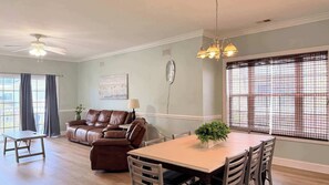 A living room with a brown leather sofa, a dining table with chairs, a ceiling fan, and a window with blinds.