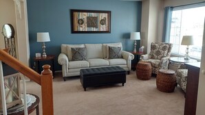 Un salon avec un mur d’accent bleu, un canapé beige, un pouf noir, un fauteuil à motifs et une table d’appoint avec une lampe.