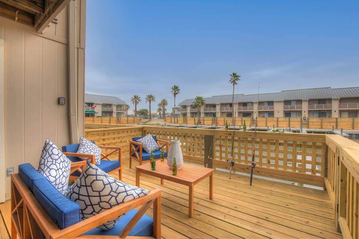 A wooden deck with blue cushioned seating and a table, overlooking a row of buildings with palm trees.
