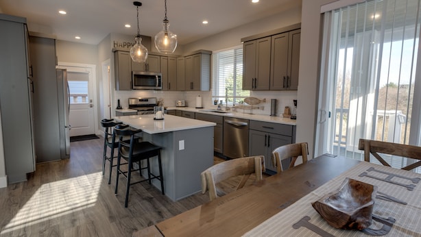 A modern kitchen with a central island, bar stools, a dining table with chairs, stainless steel appliances, and large windows. - Happiness Beach Cabin - Oyhut Bay Seaside Village (Ocean Shores)