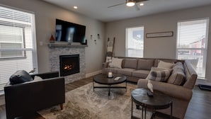 A living room with a fireplace, a sofa, a coffee table, and a television mounted above the fireplace. - Happiness Beach Cabin - Oyhut Bay Seaside Village (Ocean Shores)