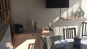A room with a television mounted on the wall, a table with a patterned tablecloth, a chair, and a small shelf with decorative items. - T2 APARTMENT IN THE CENTER OF TOWN, 5 MINUTES FROM THE SPA (Balaruc-les-Bains)