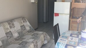 A living room with a sofa, a table with a patterned tablecloth, a refrigerator, and a wooden cabinet. - T2 APARTMENT IN THE CENTER OF TOWN, 5 MINUTES FROM THE SPA (Balaruc-les-Bains)