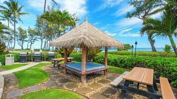 A thatched-roof gazebo with a blue cushion, surrounded by a stone pathway, palm trees, and a view of the ocean.
