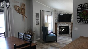 Una sala de estar con chimenea, un comedor con una mesa y sillas de madera, y un televisor montado en la pared.