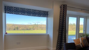 Een kamer met een groot raam, een donkere bank en uitzicht op een groen veld en bomen.