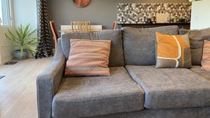 Een moderne woonkamer met een grijze bank, gestreepte kussens, een ronde wandklok en een eethoek op de achtergrond.