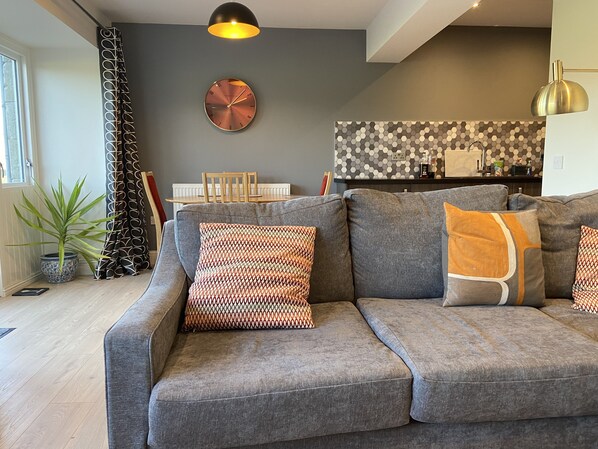 A modern living room with a grey sofa, patterned cushions, a round wall clock, and a dining area in the background.