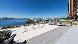 A beachside view with a wooden boardwalk, beach chairs, and a marina with boats.