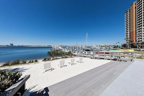 Vista de la playa con un paseo marítimo de madera, sillas de playa y un puerto deportivo con barcos.