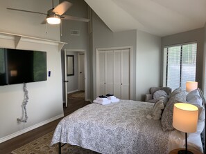 Une chambre à coucher comprenant un lit, un téléviseur, un ventilateur de plafond, une fenêtre avec des stores et une porte donnant sur une autre pièce.