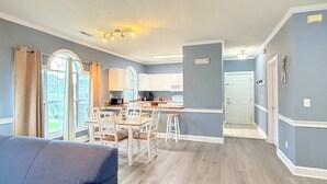 A modern kitchen with white cabinets, a dining area with wooden chairs, and a view of the outdoors through large windows. - Pond view beautiful apartment at Magnolia Place (Myrtle Beach)