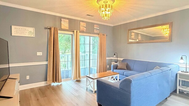A living room with a blue sofa, a wooden coffee table, a chandelier, and a large window with curtains. - Pond view beautiful apartment at Magnolia Place (Myrtle Beach)