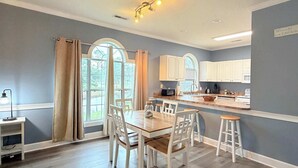 A modern kitchen with white cabinets, a dining area with a wooden table and white chairs, and a view of a garden through large windows. - Pond view beautiful apartment at Magnolia Place (Myrtle Beach)
