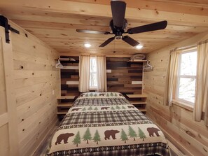 Une chambre de style cabane, dotée d’un lit, d’un ventilateur de plafond et de fenêtres avec des rideaux.