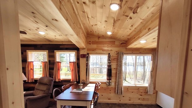 L’intérieur d’un chalet en bois comprend une table à manger, des chaises et deux fenêtres avec des rideaux.