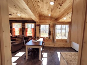L’intérieur d’un chalet en bois comprend une table à manger, des chaises et deux fenêtres avec des rideaux.