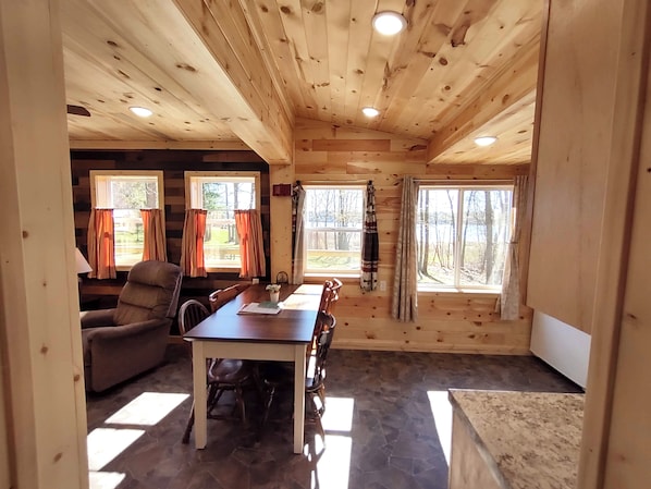 A wooden cabin interior with a dining table, chairs, and two windows with curtains.