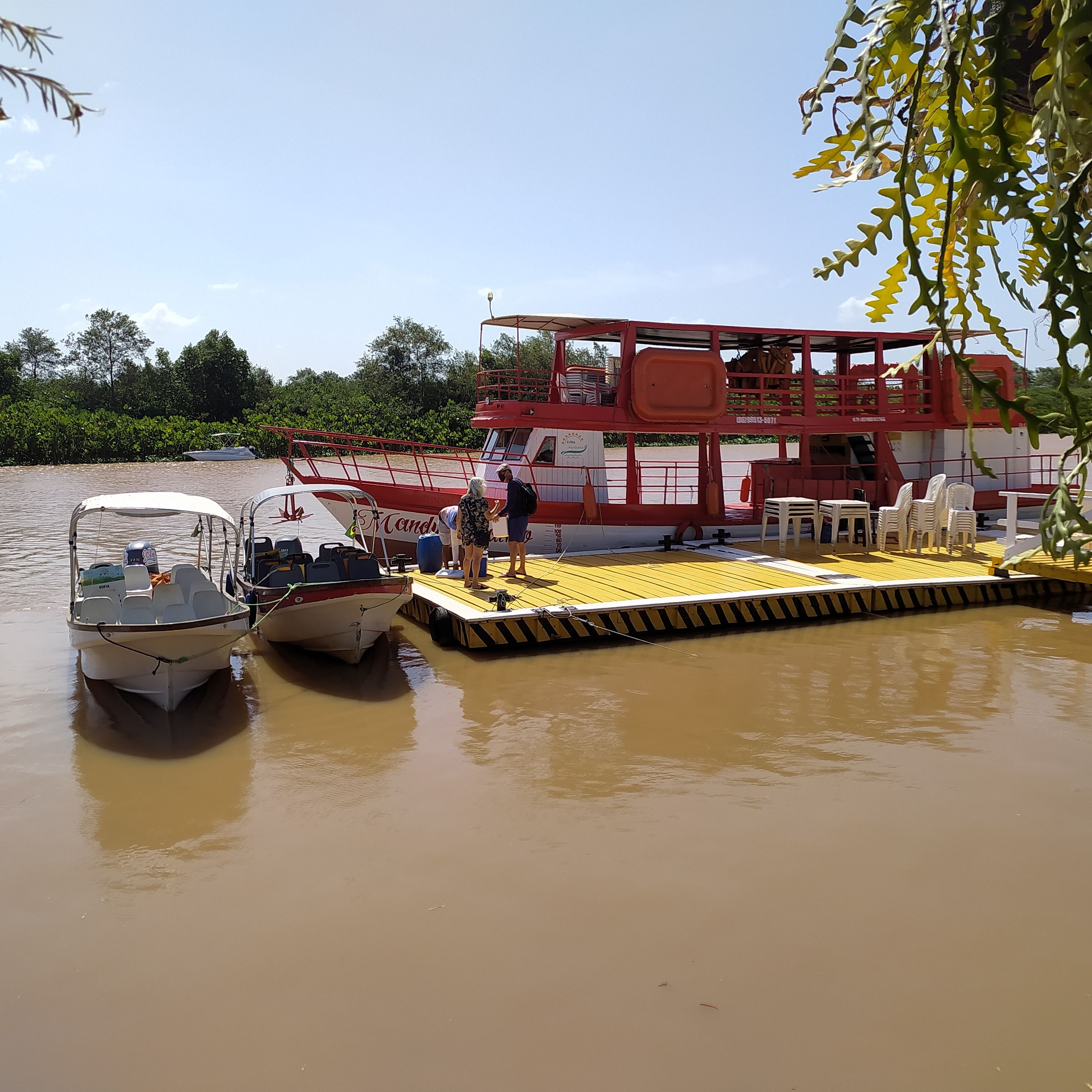 A boat docked at a pier with a red structure nearby, a small boat with a canopy, and a person standing on the pier.