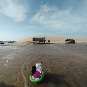 A beach scene with a child on a green inflatable ring, sand dunes, and a boat in the water. - HOTEL AVEIRO (Parnaíba)