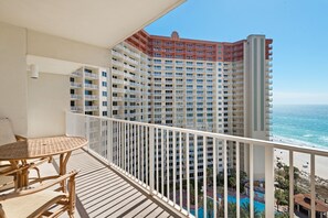 A balcony with a wicker table and chairs, overlooking a high-rise building with a pool and beach view.
