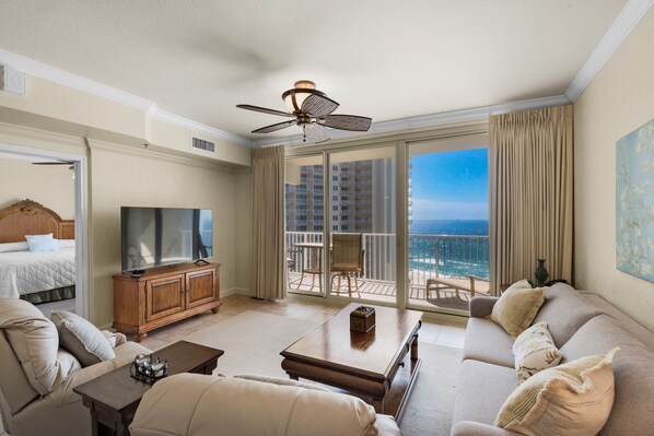 A spacious living room with a large flat-screen TV, a sofa, a coffee table, and a balcony with a view of the ocean.