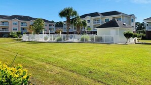 A residential area with a well-maintained lawn, a white fence, and a row of multi-story buildings with balconies. - Simply beautiful 1 floor condo in the center of MB (Myrtle Beach)
