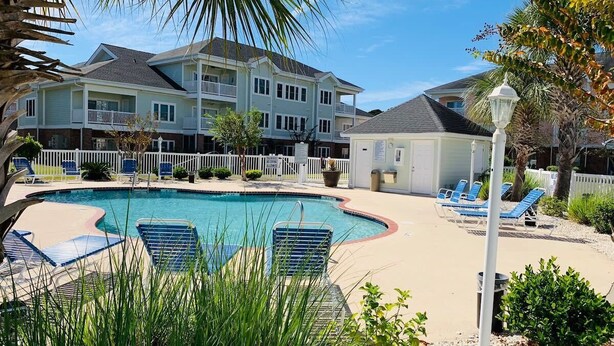 A swimming pool with lounge chairs, a fenced area, and a building with balconies in the background. - Simply beautiful 1 floor condo in the center of MB (Myrtle Beach)