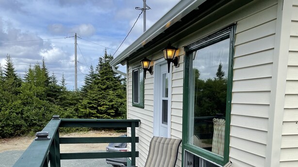 A covered outdoor seating area with two chairs and a small table, a wooden deck, and a building with a white exterior and green trim. - Hilltop Cottage (Cape Broyle)