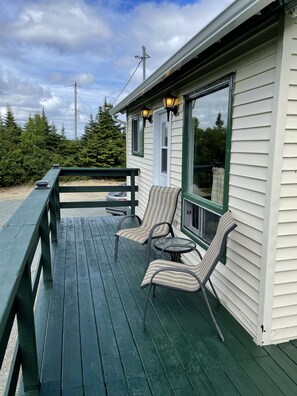 A covered outdoor seating area with two chairs and a small table, a wooden deck, and a building with a white exterior and green trim.