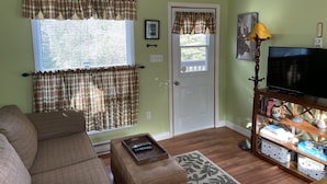 A living room with a couch, a television on a wooden stand, a door with a window, and a wall with a framed picture. - Hilltop Cottage (Cape Broyle)